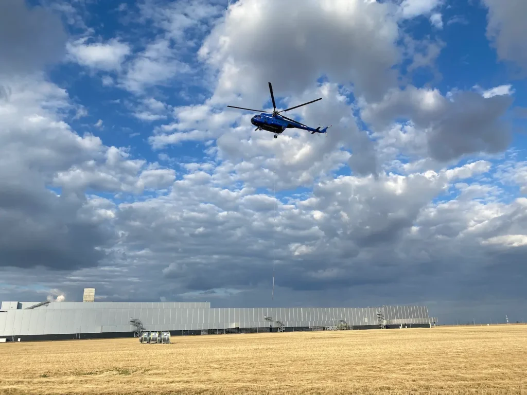 Helikopteres emelés egy ipari létesítmény mellett, háttérben felhős égbolt és üzemi terület.